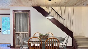 A dining area with a round table, four wooden chairs, a chandelier, and a staircase with a dark wooden handrail. - NEW Cabin by Blue Ridge Lake, Swing Bed, Firepit, Double Porches (Morganton)