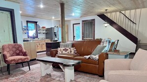 A cozy living room with a brown leather sofa, a patterned armchair, and a coffee table. A kitchen with wooden cabinets and a window is visible in the background. - NEW Cabin by Blue Ridge Lake, Swing Bed, Firepit, Double Porches (Morganton)