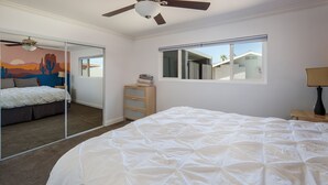 A bedroom with a large bed, a ceiling fan, a window with a view of houses, and a mirror reflecting the room. - Sunflower On the Lake (Lake Havasu City)
