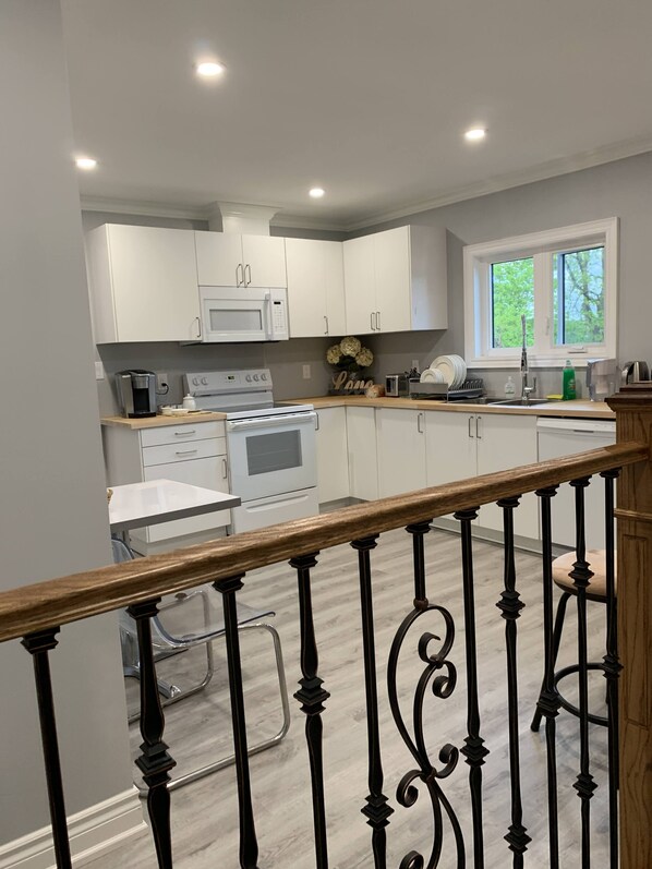 A modern kitchen with white cabinets, a built-in oven, microwave, and coffee maker. There is a window above the sink, and a wooden railing in the foreground.