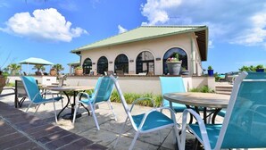An outdoor patio with blue chairs and tables, a building with arched windows, and a clear blue sky.