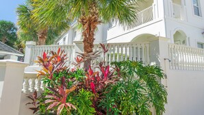 Ein weißes, zweistöckiges Haus mit Balkon, umgeben von üppiger Vegetation, darunter eine Palme und verschiedene Pflanzen.
