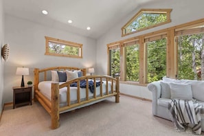 Quarto com cama de madeira, mesa de cabeceira, luminária, janela com vista para árvores e sofá branco.