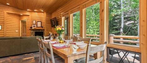 A wooden cabin interior with a dining table set for four, a sofa, and large windows offering a view of the forest.