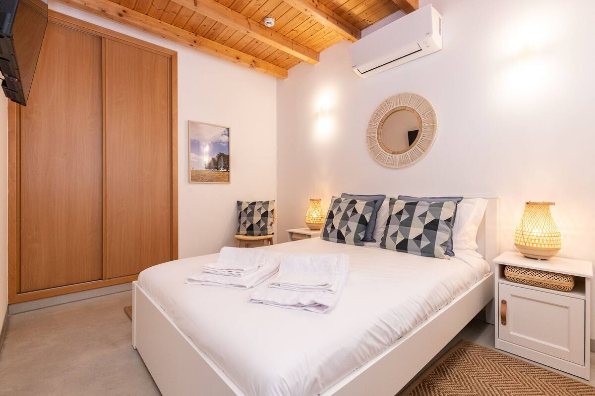 A bedroom with a large bed, wooden wardrobe, bedside tables with lamps, and a wall-mounted air conditioner.