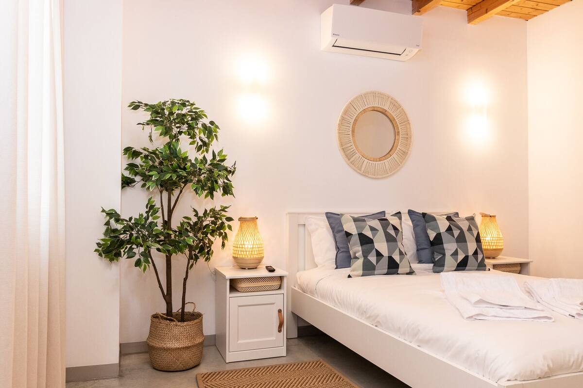 A bedroom with a bed, bedside lamps, a wall-mounted air conditioner, a mirror, and a potted plant.