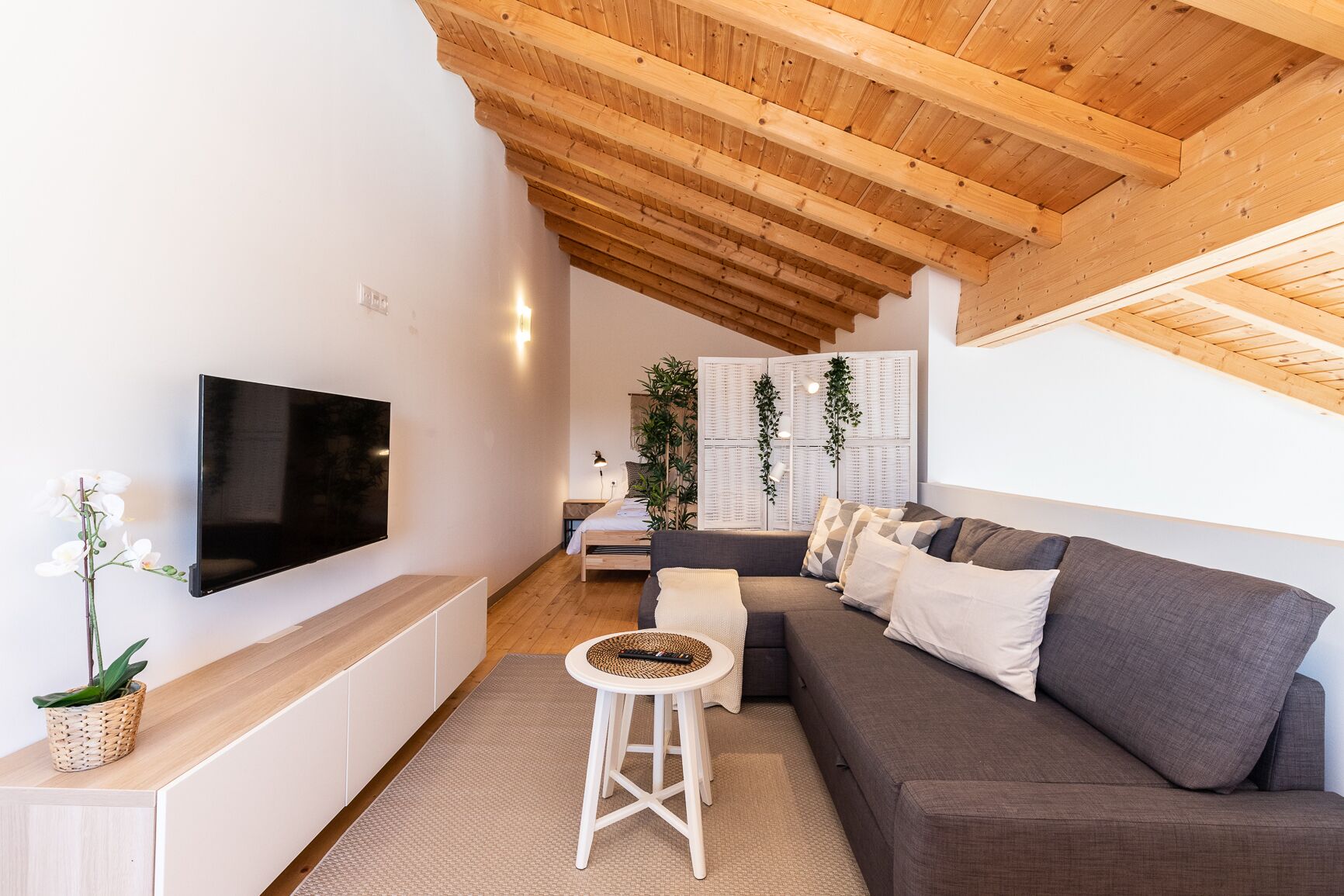 A modern living room with a flat-screen TV, a sofa, a small round table, and a wooden ceiling.
