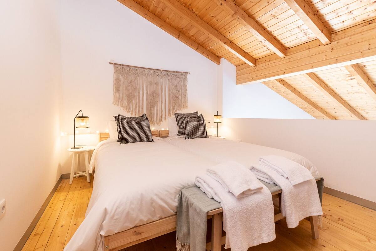 A bedroom with a large bed, wooden beams, a hanging macrame curtain, and bedside lamps.