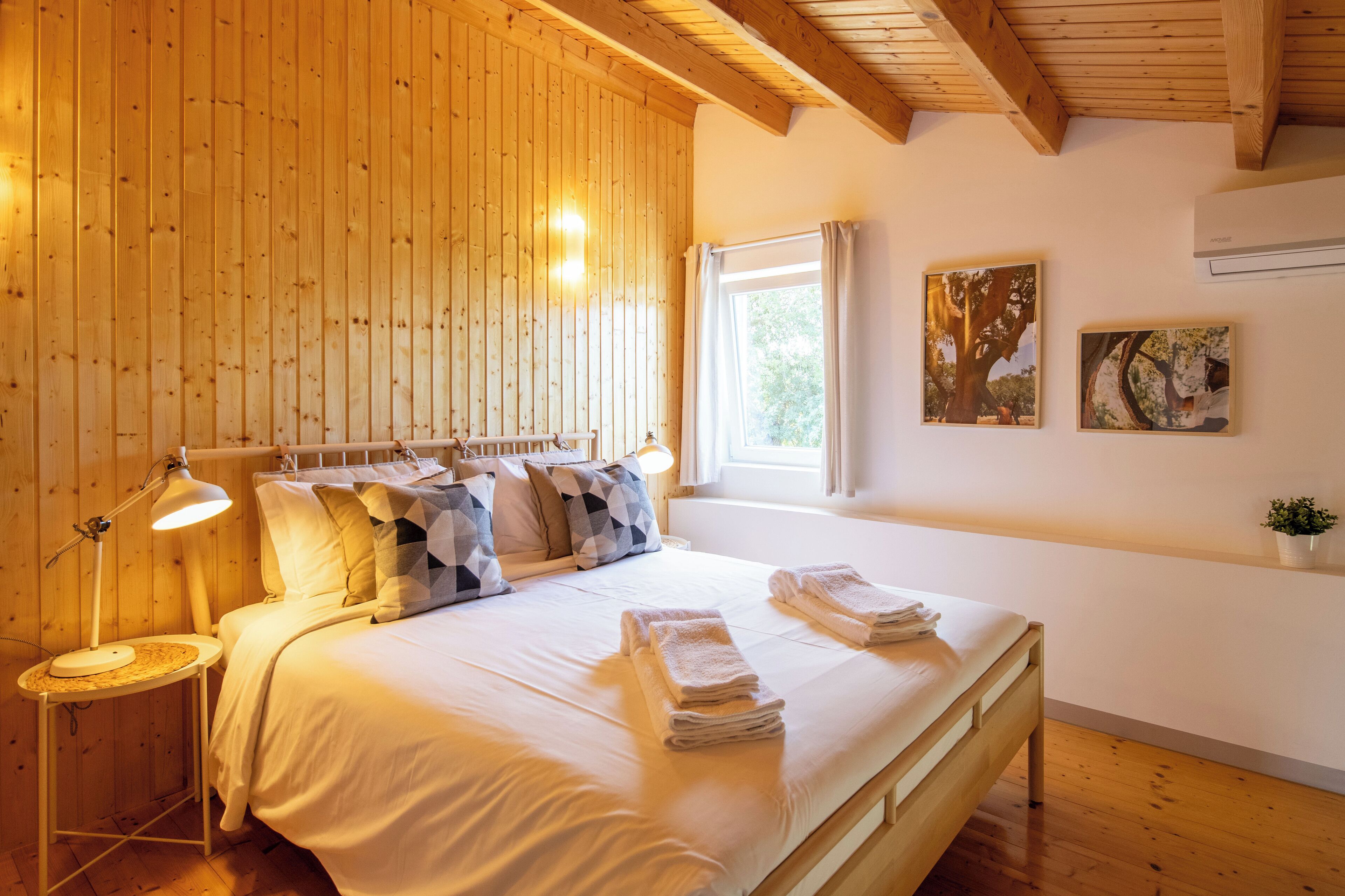 A bedroom with a wooden ceiling, a bed with white linens, a bedside lamp, and two framed pictures on the wall.