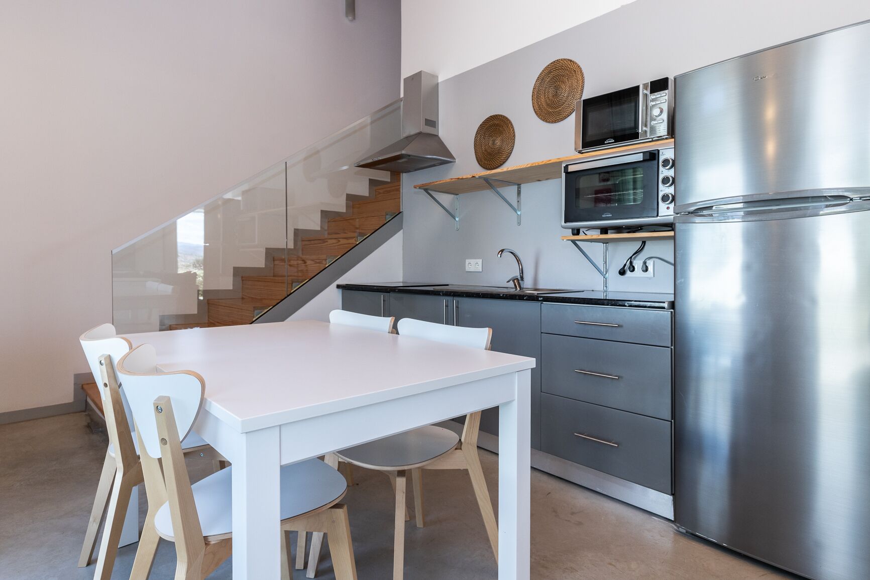 Cocina moderna con refrigerador de acero inoxidable, microondas y estufa. Hay una mesa de comedor blanca con sillas de madera. Una escalera con barandales de vidrio conduce a otro nivel.