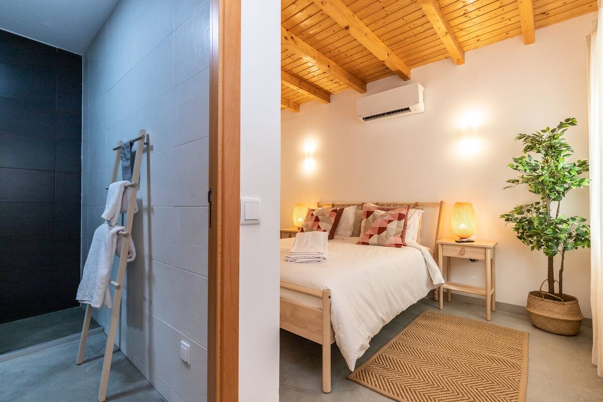 A bedroom with a wooden ceiling, a bed with patterned pillows, a bedside lamp, and a potted plant.