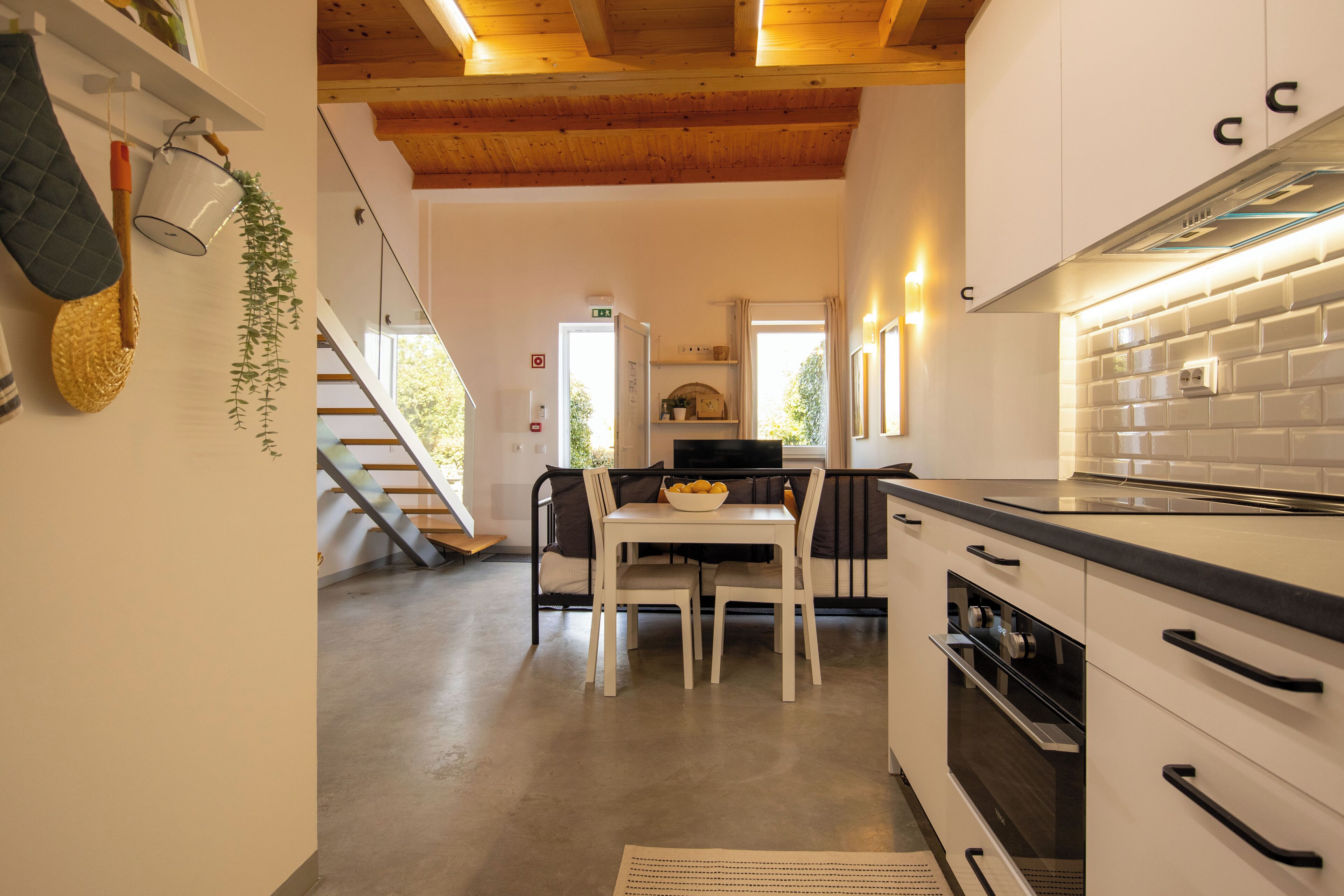 Cuina moderna amb armaris blancs, tauleig negre i sostre de fusta. Hi ha una zona de menjador amb taula i cadires, i una escala que condueix a un altre pis.
