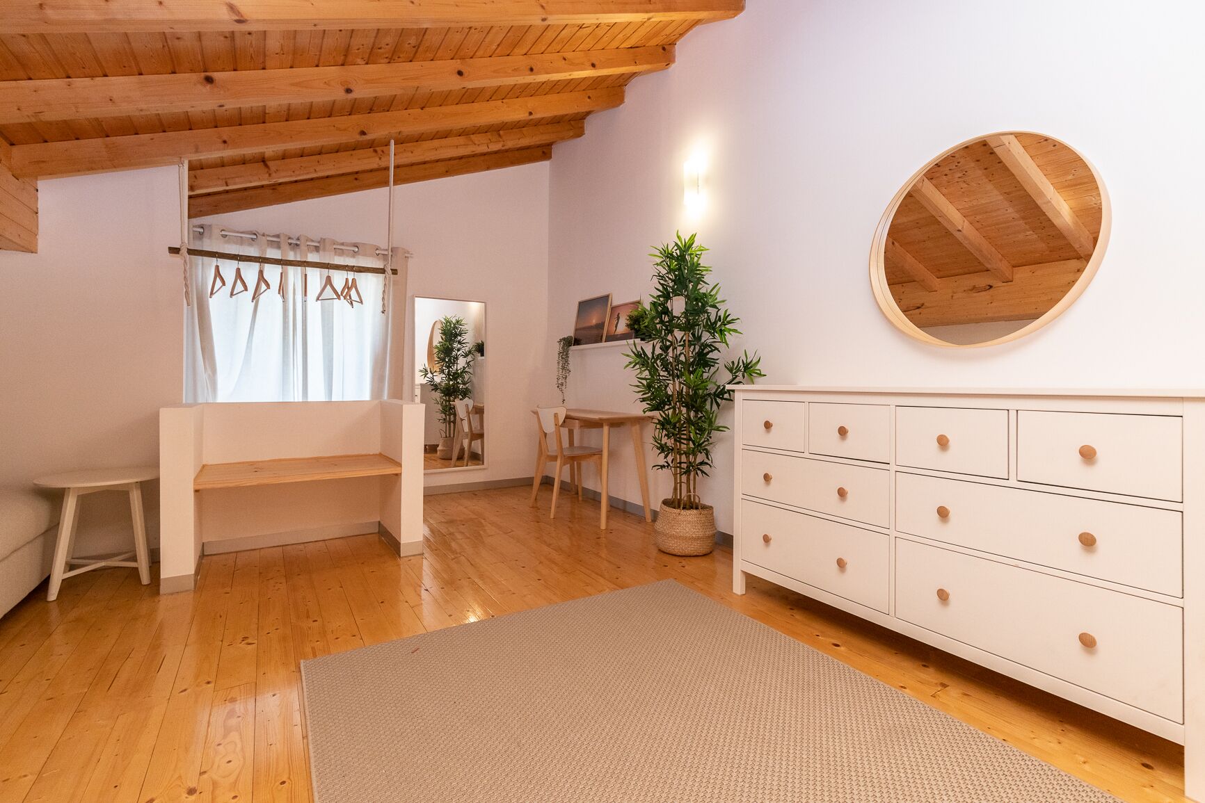 Ein Zimmer mit Holzboden, einer weißen Kommode mit Schubladen, einem runden Spiegel, einem kleinen Tisch, einer Bank und zwei Zimmerpflanzen.