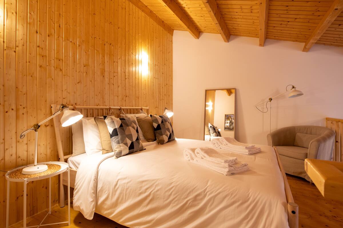 A cozy bedroom with a large bed, two bedside lamps, a chair, and a wooden ceiling.