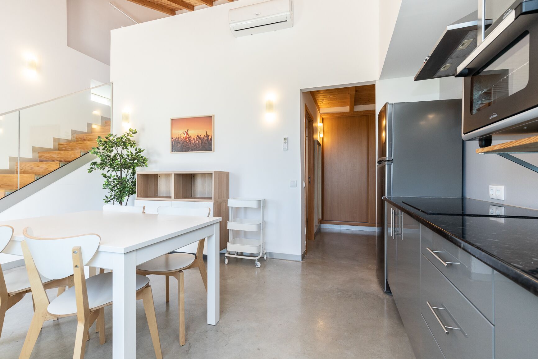 A modern kitchen with a black countertop and stainless steel appliances, a dining area with a white table and wooden chairs, and a staircase with glass railings.
