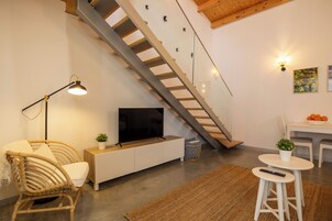 Una sala de estar moderna con un sofá, un televisor sobre un mueble blanco, una escalera, una mesa de comedor con sillas y una planta pequeña sobre la mesa.