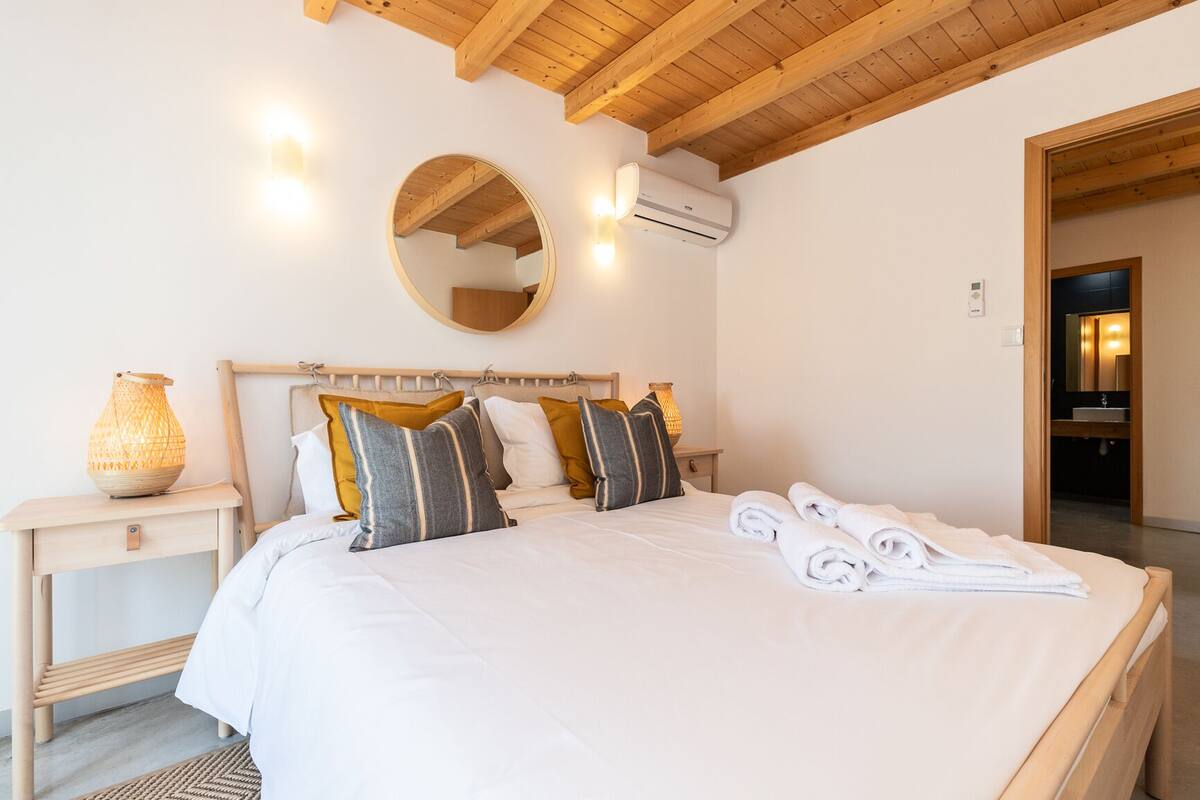 A bedroom with a wooden ceiling, a round mirror, a bed with white bedding and striped pillows, a bedside lamp, and a folded towel on the bed.