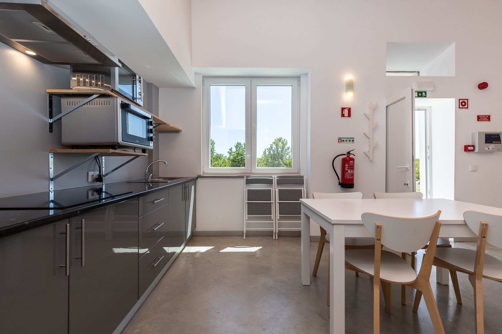 A modern kitchen with a large window, a dining area with a white table and chairs, and a fire extinguisher mounted on the wall.
