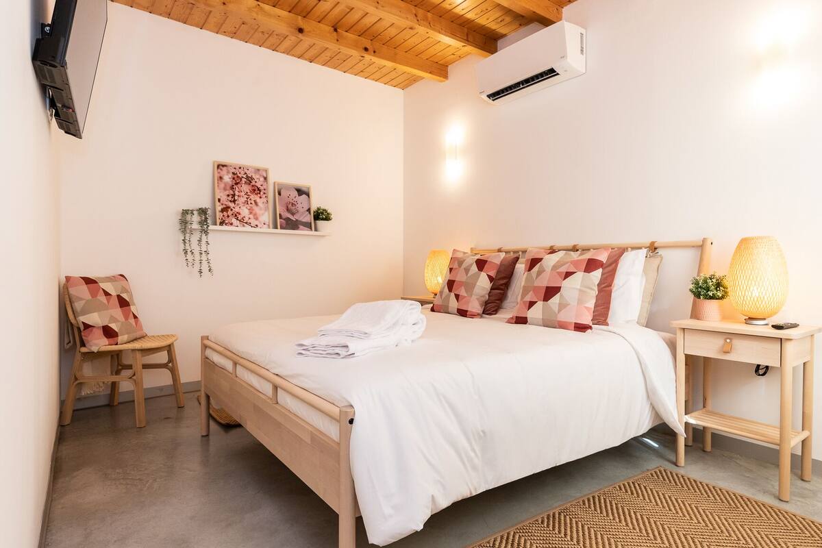 A bedroom with a wooden bed, a bedside table with a lamp, a chair, and wall-mounted air conditioning.