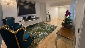 A modern living room with a large flat-screen TV, a desk with an Apple iMac, a teal armchair, a wooden coffee table, and a green patterned rug. - Pier House Accommodation (Funchal)