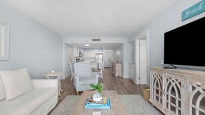A living room with a sofa, a coffee table with books and a plant, a TV on a cabinet, and a kitchen in the background. - Beachcrest Gulf-Front Private Beach- Chair & umbrella service -March-Oct (Santa Rosa Beach)