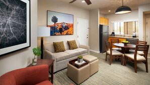 A modern living room with a sofa, ottoman, and red armchair. There is a dining area with a table and chairs. A kitchen is visible in the background with a refrigerator and a window. - Westin Desert Willow Resort (Palm Desert)