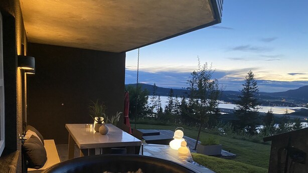 Un balcon avec vue sur un lac et des montagnes, une table avec un vase et une bougie, et une balançoire suspendue.