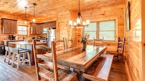 L’intérieur d’un chalet en bois comprend une grande table à manger, des chaises en bois, un coin cuisine et un lustre.