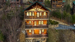 Une cabane en rondins à plusieurs étages, dotée d’un balcon au deuxième étage, entourée d’arbres et bordée d’un sentier en pierre.