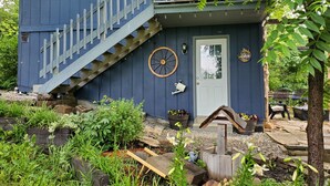 Una casa azul con escalera de madera que lleva a una terraza, rodeada de follaje y plantas en macetas.