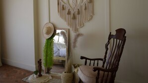 Une pièce avec un plancher en bois, un fauteuil à bascule, une décoration murale en macramé et une petite plante.
