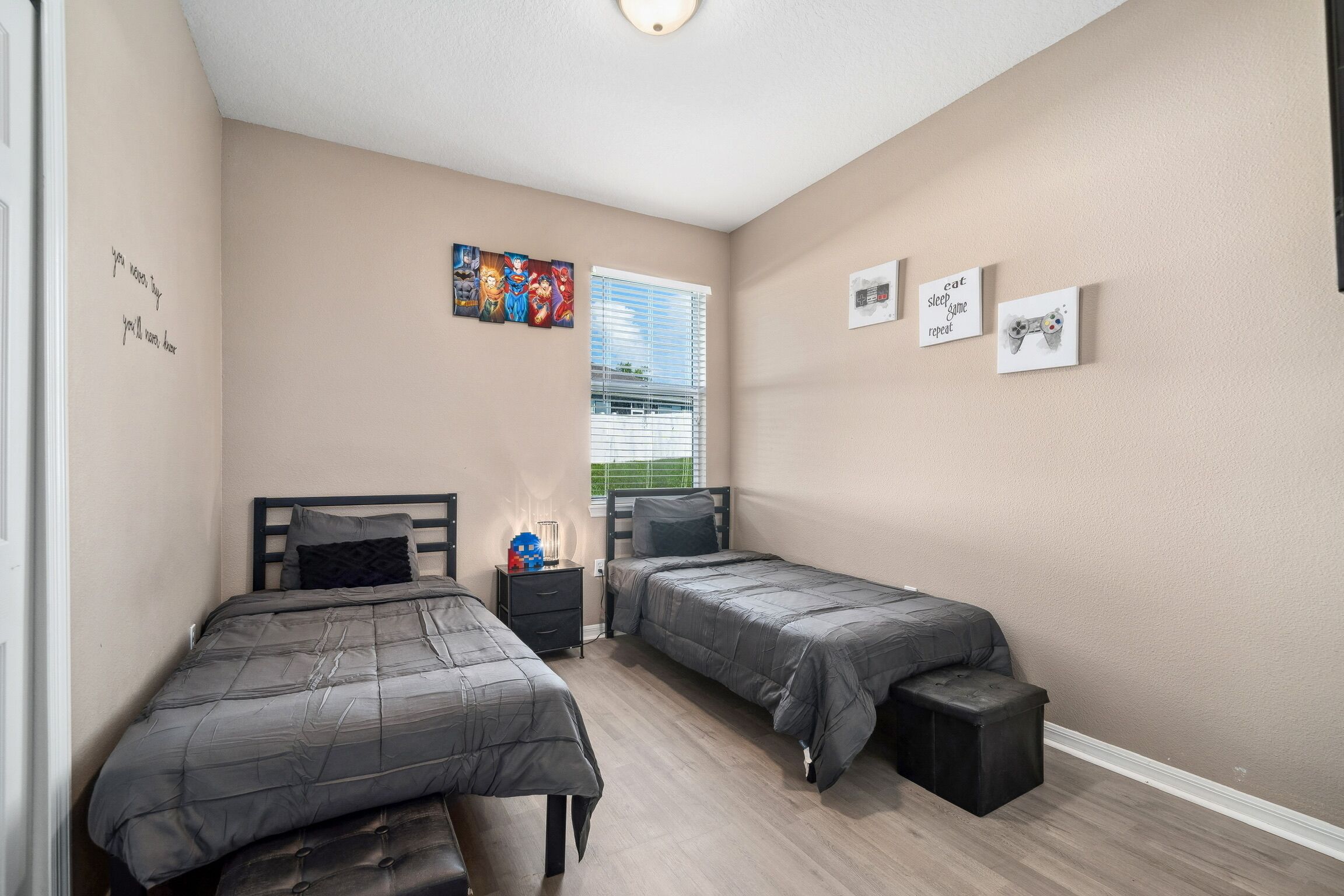 A bedroom with two beds, a window with blinds, a bedside table with a lamp, and wall art.