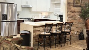 Una cocina moderna con armarios blancos, un refrigerador de acero inoxidable, una estufa a gas y una pared de acento de piedra. Hay tres taburetes de bar con asientos acolchados y estructura de metal.