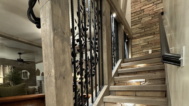 Una escalera con detalles de madera y barandillas de metal decorativo, que conduce a una habitación con pared de piedra y vistas al exterior.