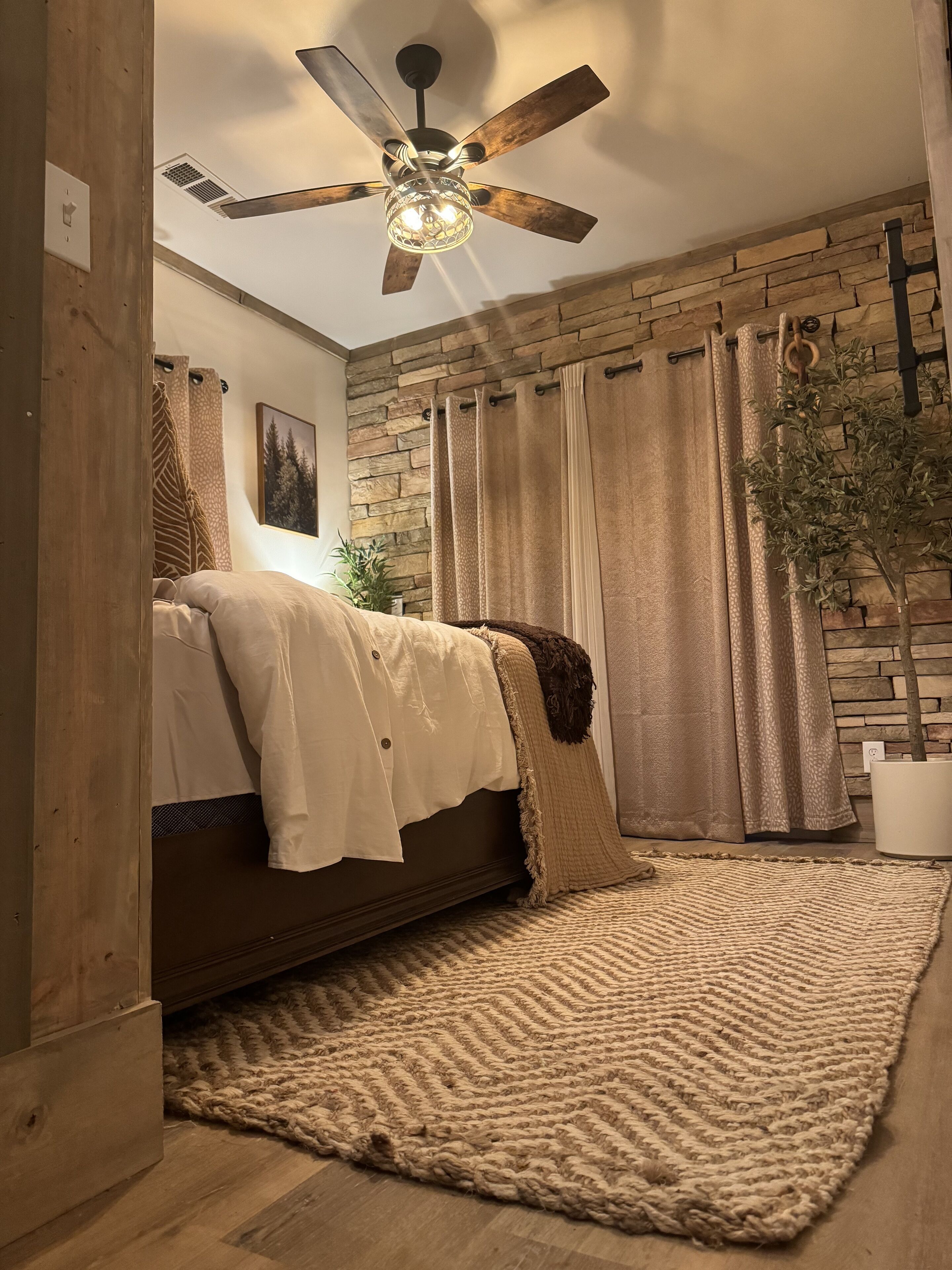 A bedroom with a bed, a ceiling fan, a stone wall, and a window with curtains.