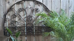 Una cerca de madera con un portón metálico decorativo, plantas en macetas, un farol y un banco blanco.