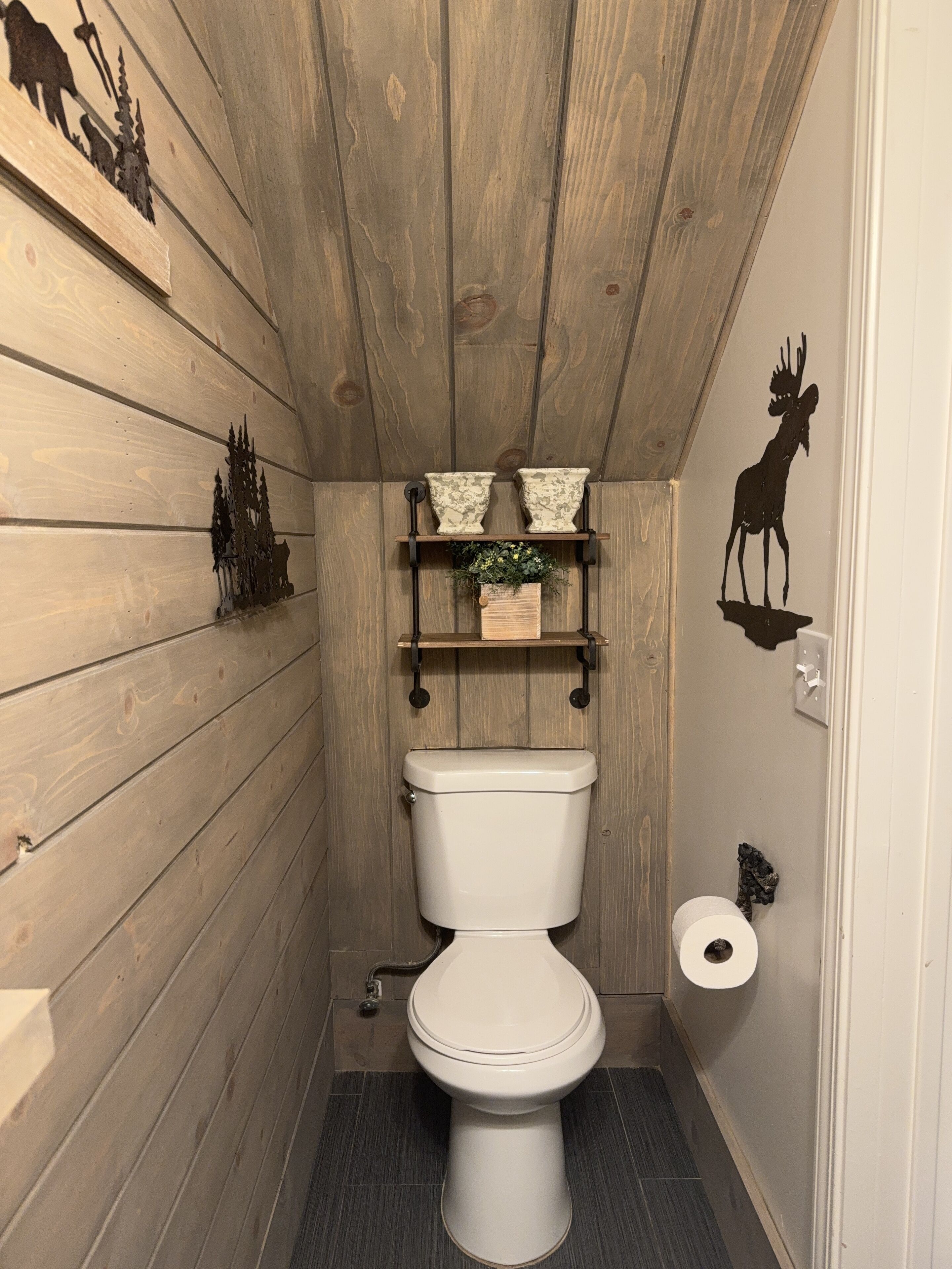 A small, rustic bathroom with wooden walls, a white toilet, a shelf with decorative items, and a deer decal on the wall.