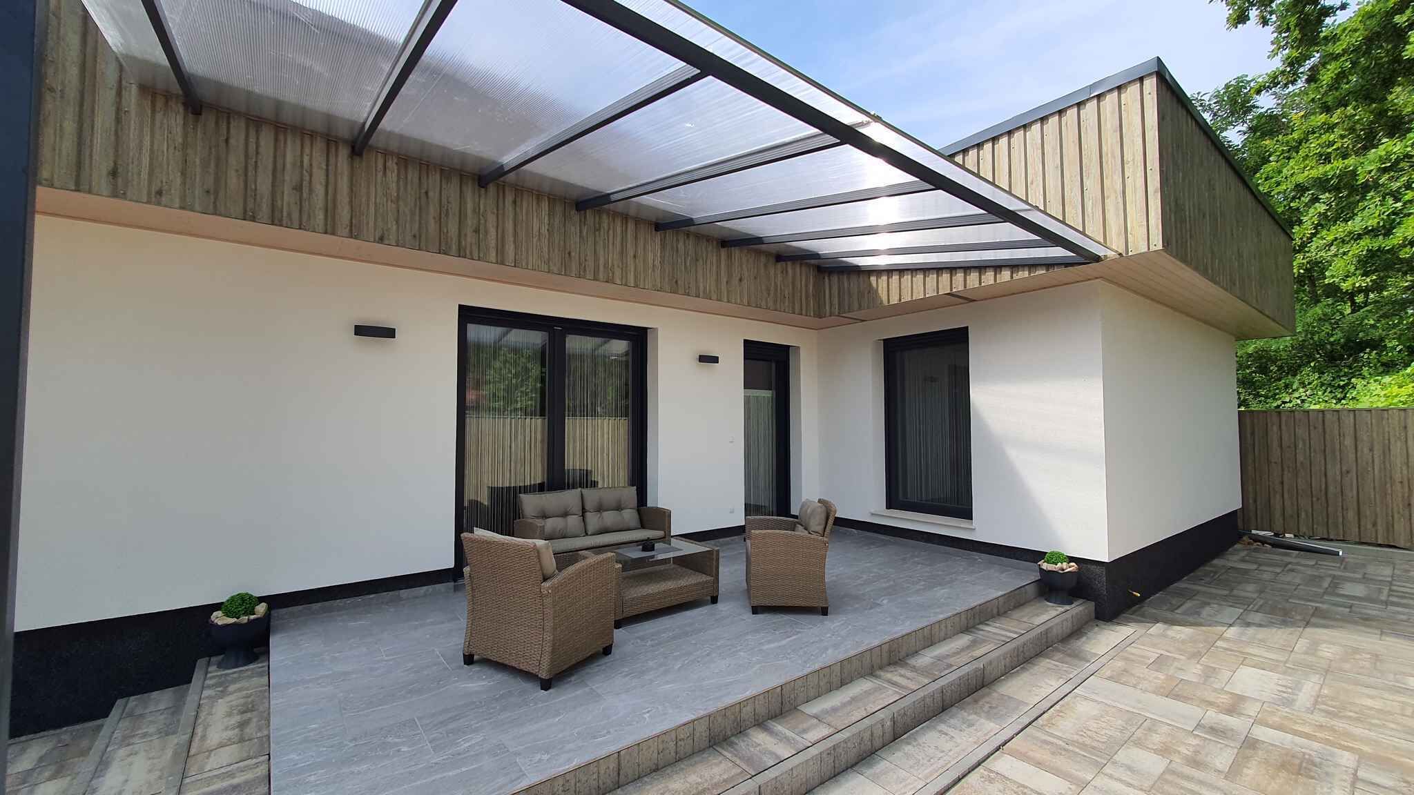 Un patio moderno con cerramiento de vidrio, mobiliario exterior y una terraza de madera.