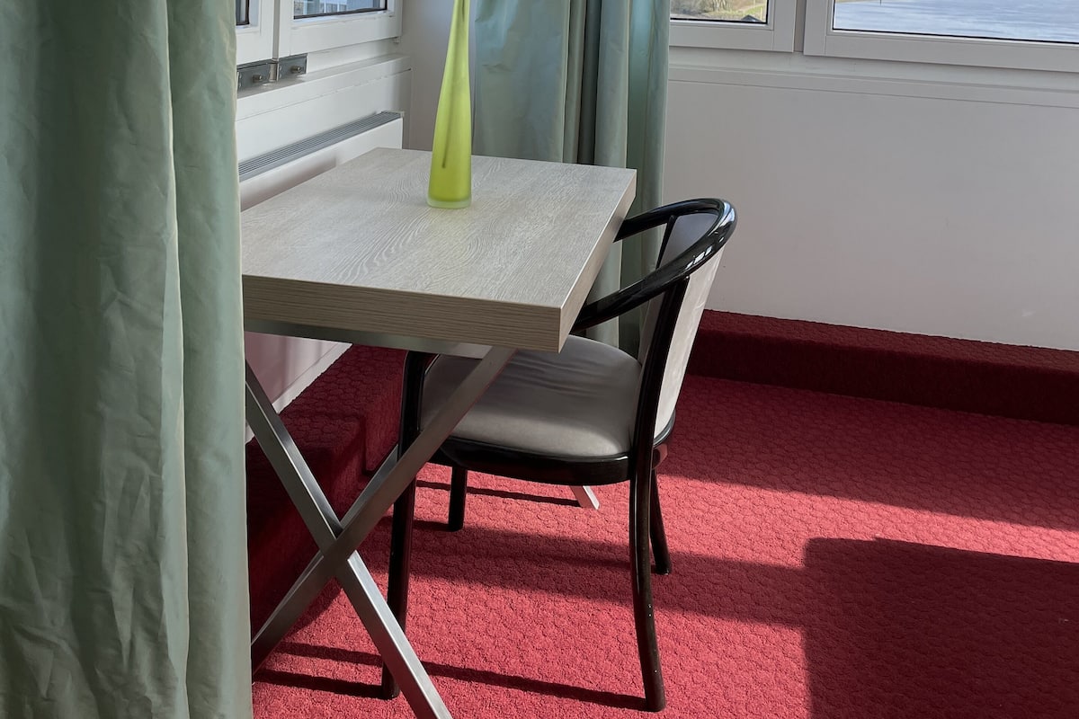 A room with a table, a chair, a vase with flowers, and a view of a body of water through large windows.