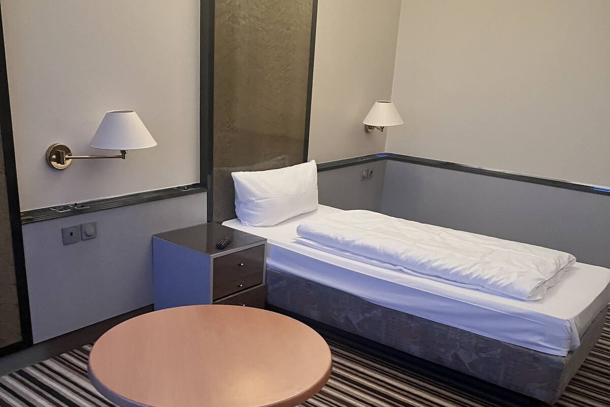 A hotel room with a bed, two bedside lamps, a round table, a small nightstand, and a woven chair.