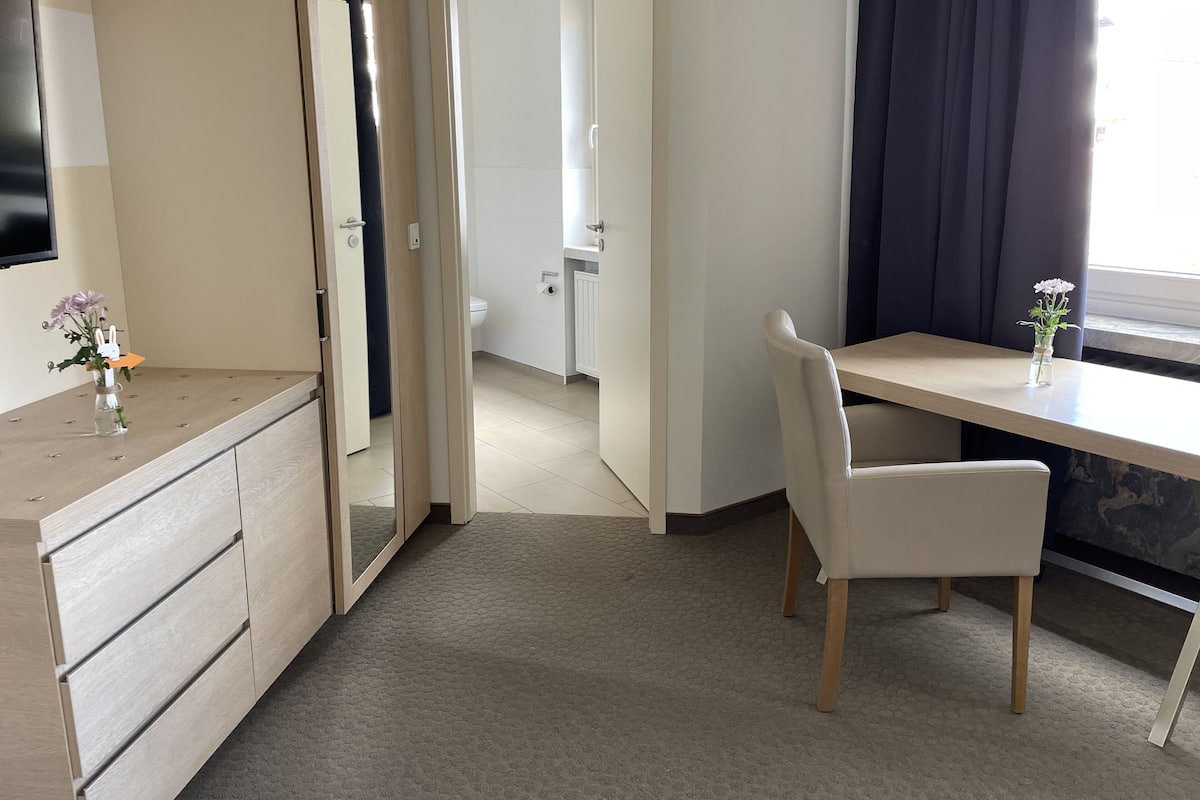 A modern bedroom with a wooden dresser, a desk, and a chair. There is a mirror on the dresser, and a vase with flowers on the desk. The room has a tiled floor and a window with curtains.