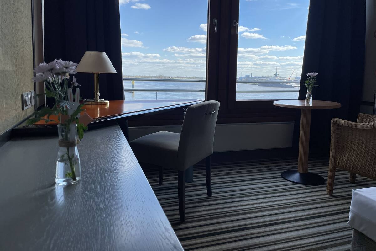 A hotel room with a large window, a desk with a lamp, a chair, a small table with a vase of flowers, and a bed.