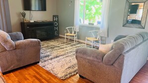 A living room with two grey sofas, a wooden floor, a large rug, a TV on the wall, and a window with curtains. - Cheerful 4-Bedroom In Beautiful Dandridge  (Dandridge)