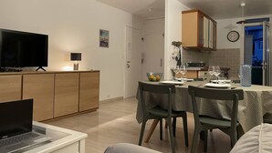 Une cuisine moderne avec des meubles en bois, un coin repas avec deux chaises et une table dressée pour deux personnes, un téléviseur posé sur un meuble et un ordinateur portable sur un bureau.