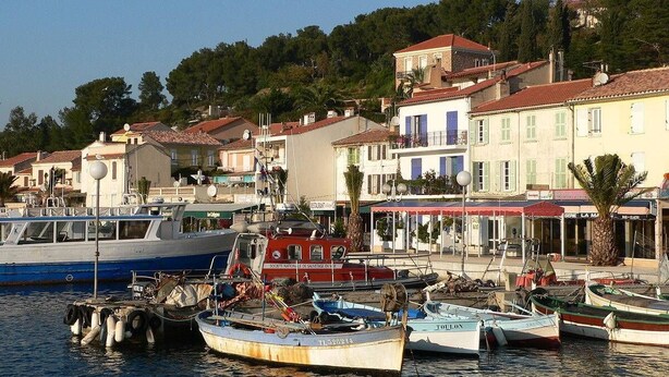Un port de plaisance où sont amarrés plusieurs bateaux, entouré de bâtiments aux toits recouverts de tuiles rouges, avec une colline en arri ère-plan.