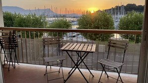 Un balcon avec une table en bois et deux chaises, donnant sur une marina où sont amarrés des bateaux, et offrant une vue sur le coucher du soleil.