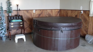 A room with a wooden wall, a small wooden cabinet with drawers, a white stool, a large brown hot tub, and a clock on the wall. - 3 br country home close to town with hot tub (Muncie)