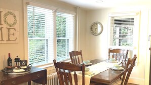 A dining room with a wooden table, six chairs, a clock, and a window with blinds. - Quarry Landing - Center of Town & River View - 3 Bedroom (Port Deposit)