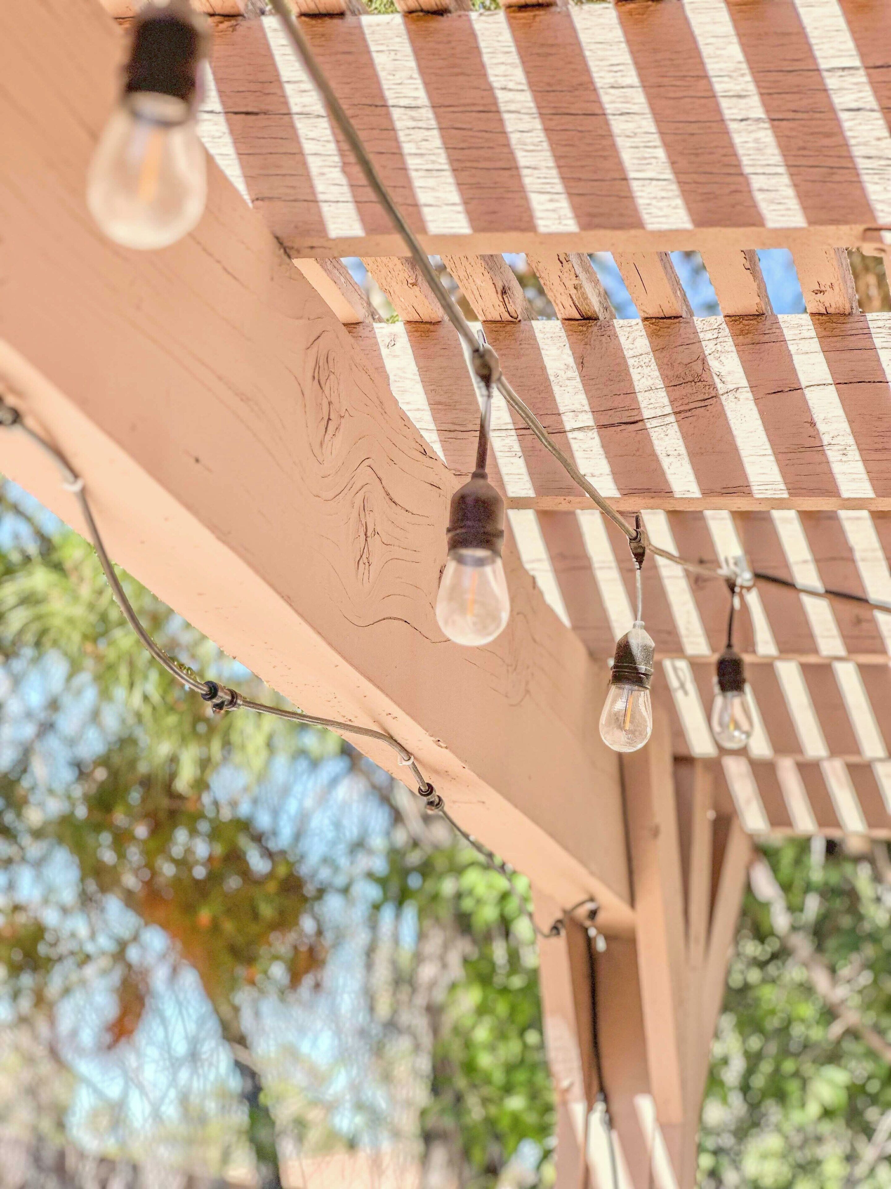 A wooden pergola with a striped pattern and hanging light bulbs.