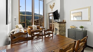 A dining area with a wooden table and chairs, a stone fireplace, and a large window with a view of a forested mountain. - Spacious 3bdrm Family Retreat Ski in/out w/hot tub (Sun Peaks)